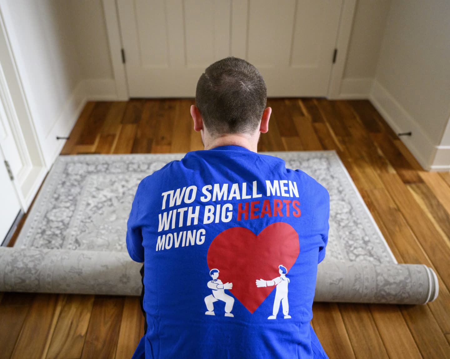 Man in a Two Small Men with Big Hearts Moving shirt rolling up a rug on a wooden floor.