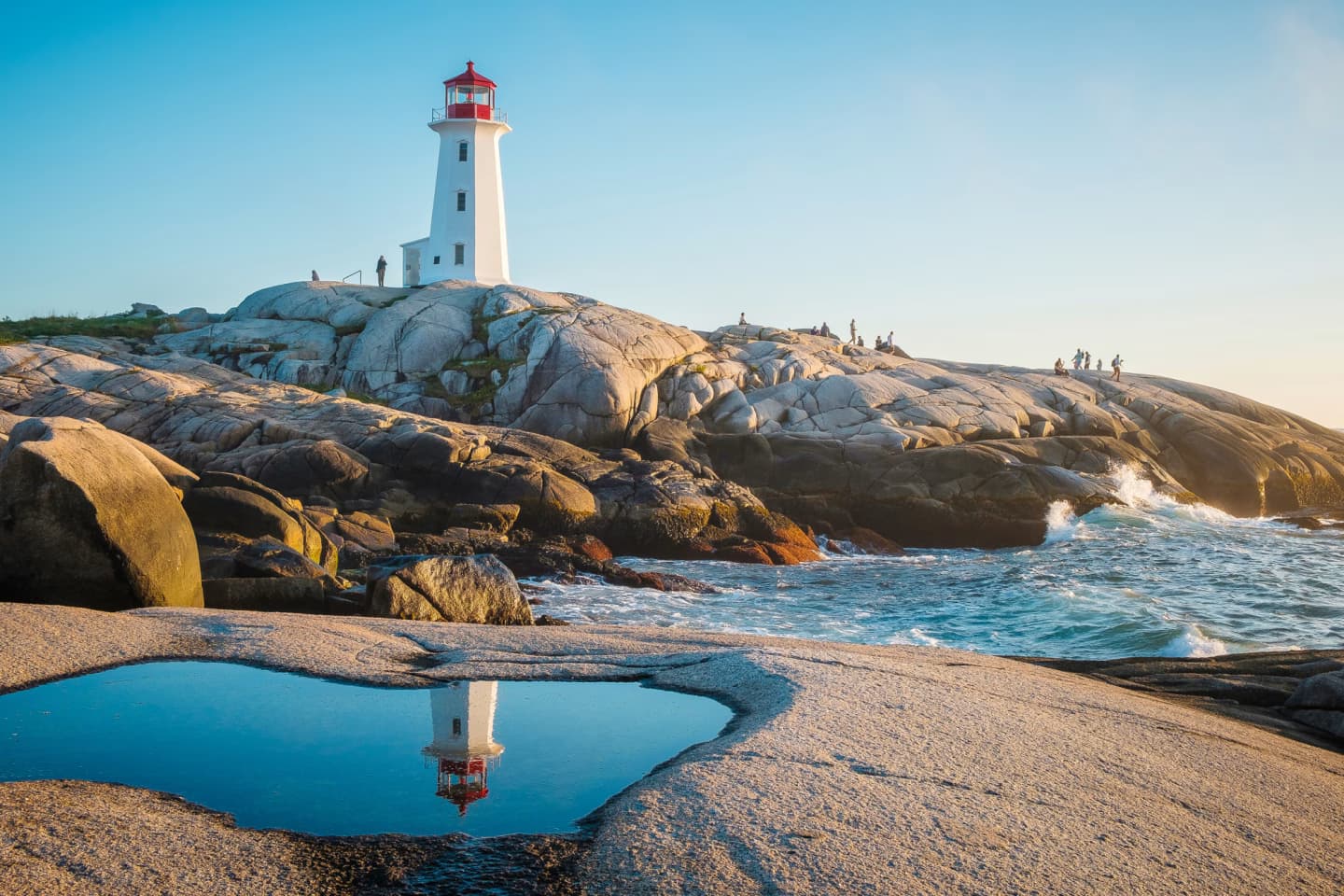 Peggy's Cove Lighthouse in Nova Scotia reflected in a tidal pool on a rocky shoreline during sunset.