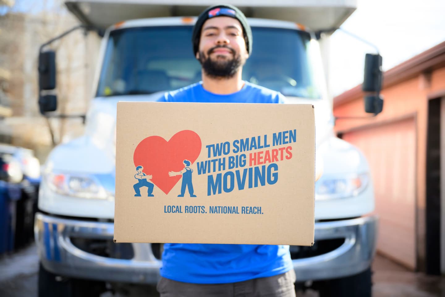 A mover in a "Two Small Men with Big Hearts" shirt is holding a cardboard box with the company logo and slogan "Local Roots, National Reach," standing in front of a moving truck.