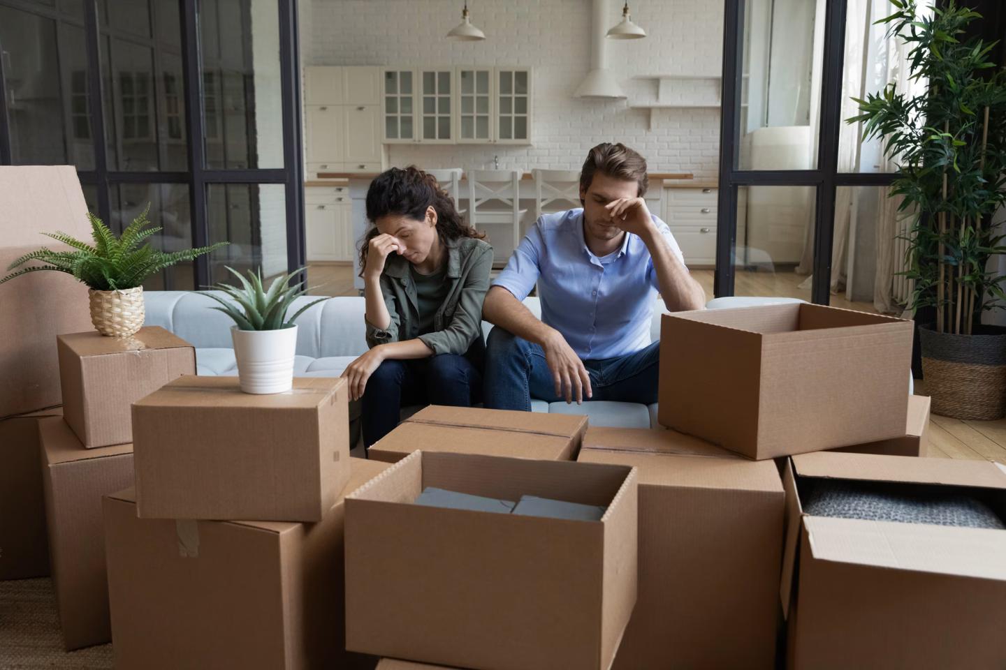 Stressed couple sitting on a couch surrounded by moving boxes in a new home.