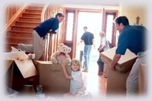 A family moving into a new home. A child sits on a moving box with a teddy bear, while two movers carry boxes. Another person is seen entering the house through the front door.