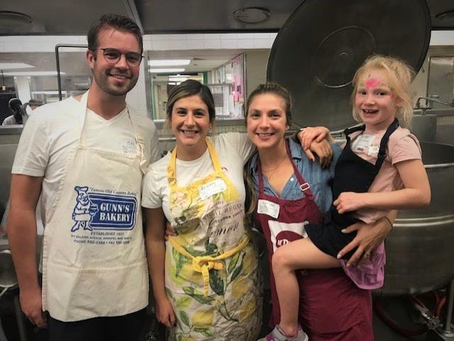 Four people are posing in a kitchen, with three adults wearing aprons and a young child in an apron being held. They are all smiling, and large cooking pots are visible in the background.