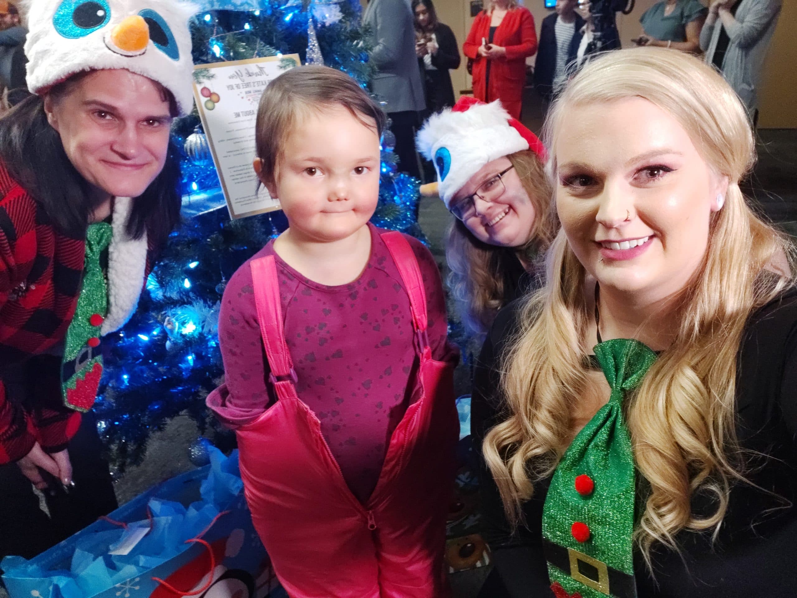 Group of people, including a child, posing in front of a decorated Christmas tree at a holiday event.