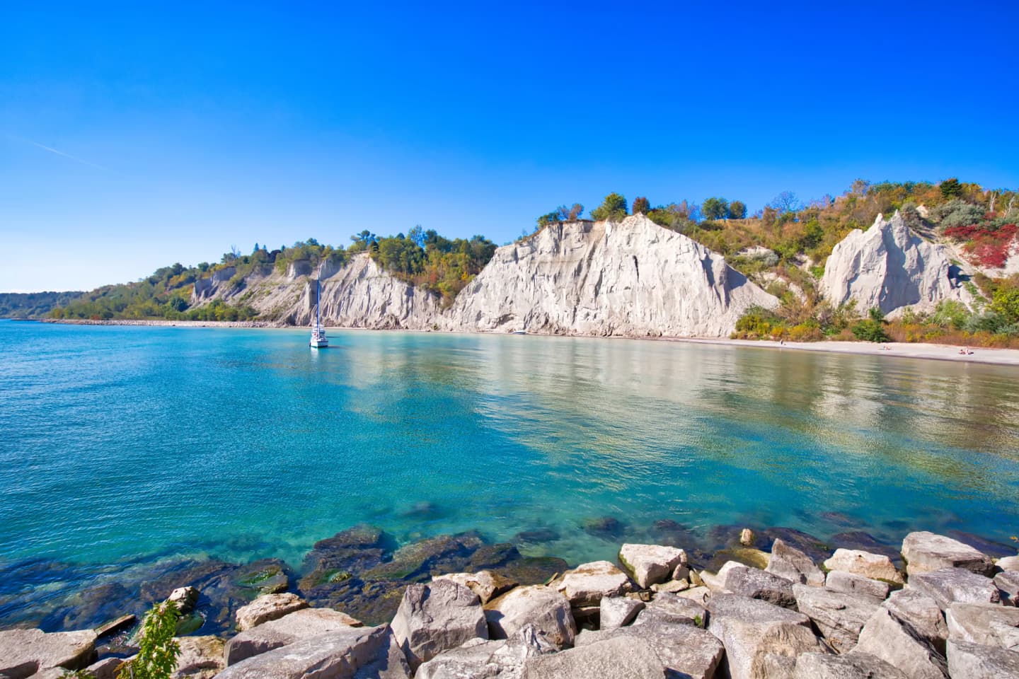 Scarborough Bluffs in Toronto, Ontario, with clear blue water, rocky shoreline, and a sailboat on a sunny day.