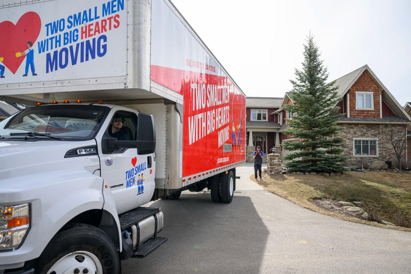 Two Small Men with Big Hearts Moving truck being backed up towards a house, with one mover guiding the truck and another in the driver's seat.