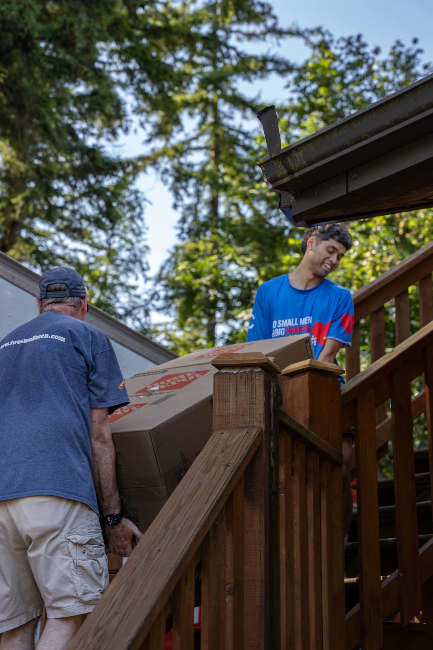 Two movers are carrying a large cardboard box up a wooden staircase, with one mover wearing a "Two Small Men with Big Hearts" shirt. Trees and a house are visible in the background.