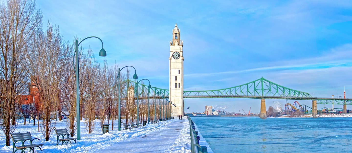 Clock tower along a snowy riverside path with the Jacques Cartier Bridge in the background.