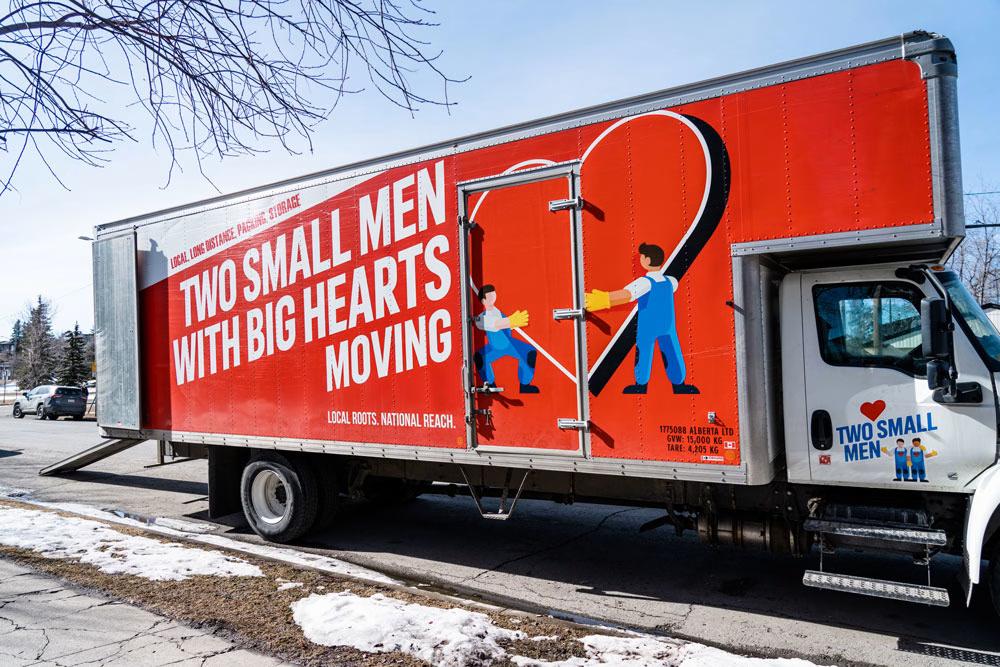 A "Two Small Men with Big Hearts Moving" truck is parked on a street during winter, featuring the company’s logo with cartoon movers and a large heart on its red side. Snow is visible on the ground.