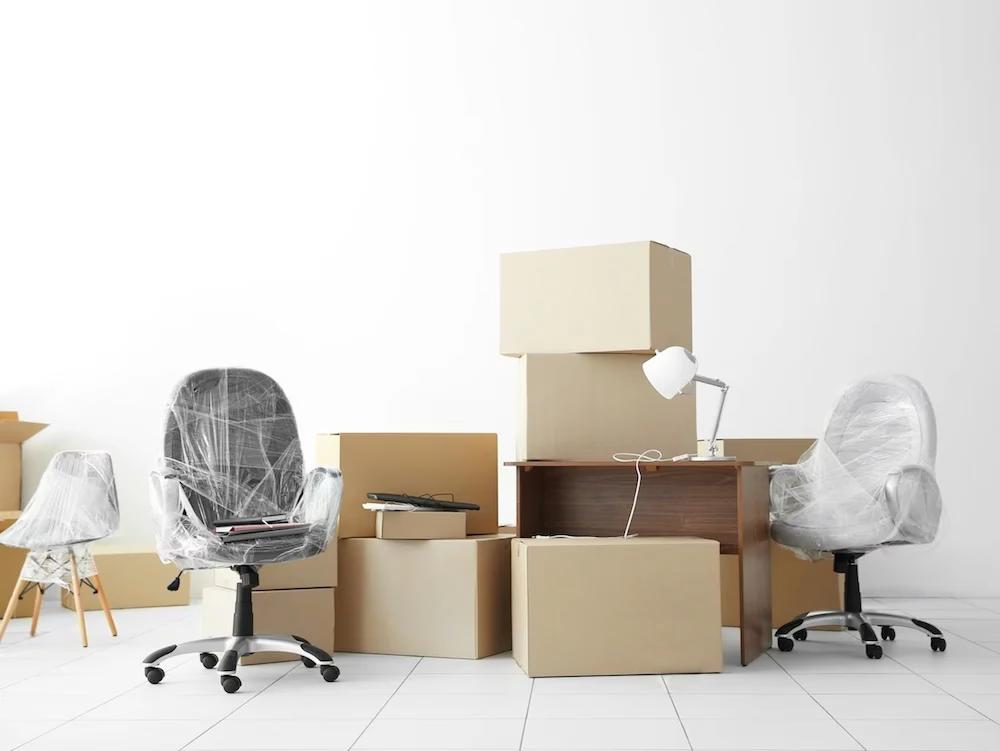 An office space in transition, filled with packed moving boxes, wrapped chairs, and furniture ready for relocation.