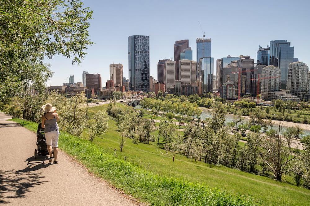 Person pushing a stroller along a path with the Calgary skyline in the background on a sunny day.