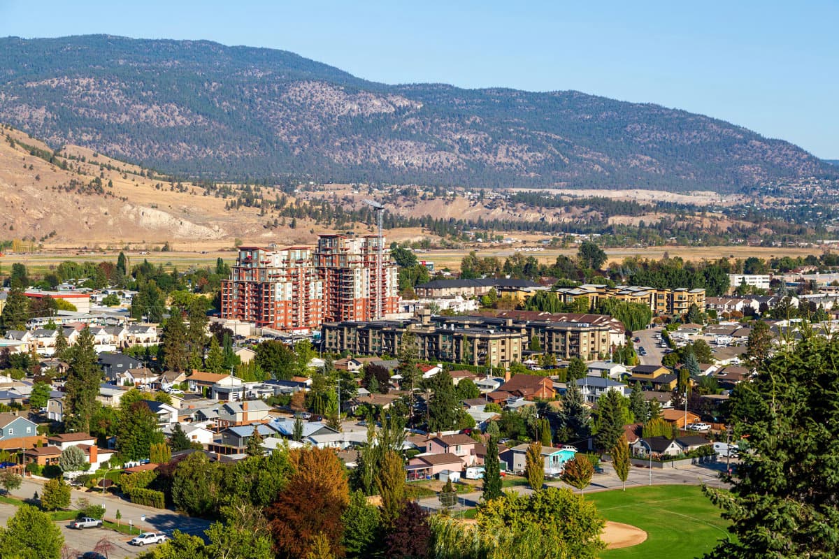 Aerial view of Penticton with residential buildings, greenery, and mountains in the background on a sunny day.