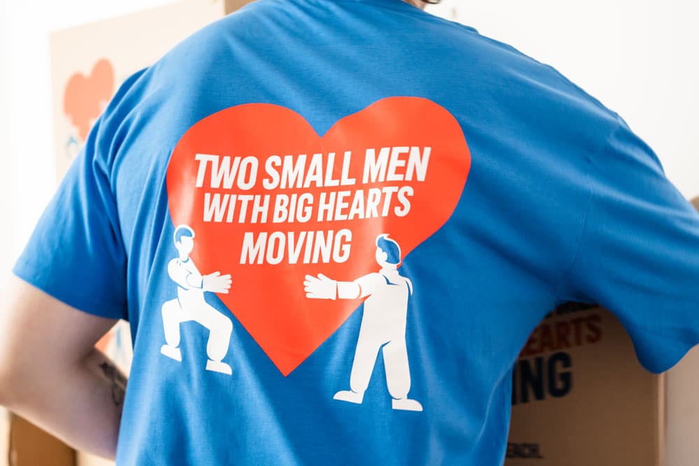 A mover is wearing a blue "Two Small Men with Big Hearts Moving" shirt with the company logo and a large red heart on the back, standing in front of cardboard boxes.