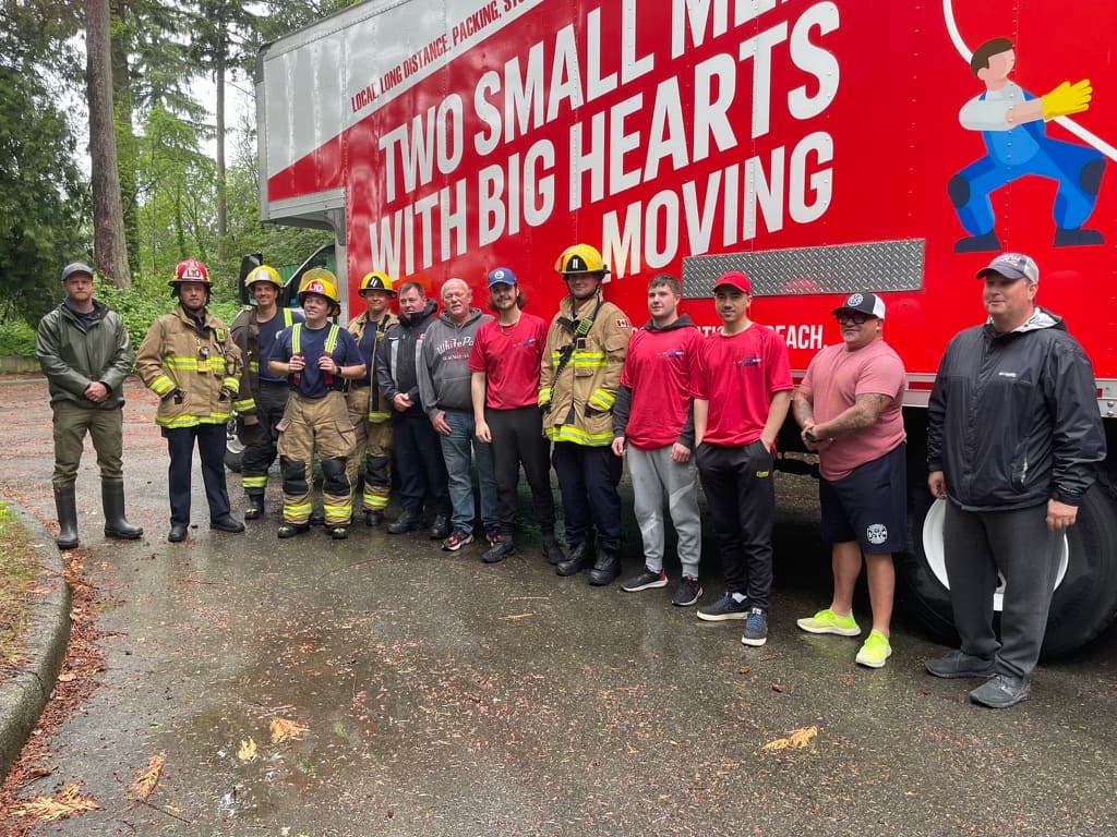 Group of firefighters and movers standing in front of a Two Small Men with Big Hearts Moving truck.