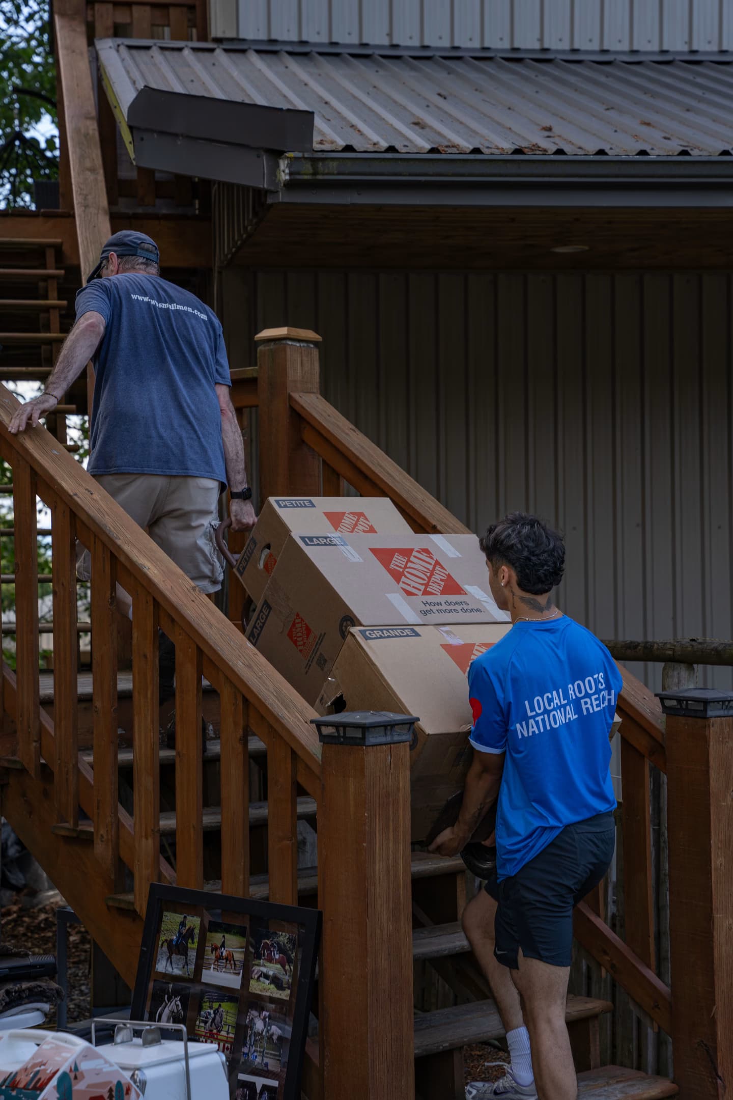 Two movers are carrying large boxes up a wooden staircase, one wearing a "Two Small Men with Big Hearts" shirt. A house with a metal roof is visible in the background.