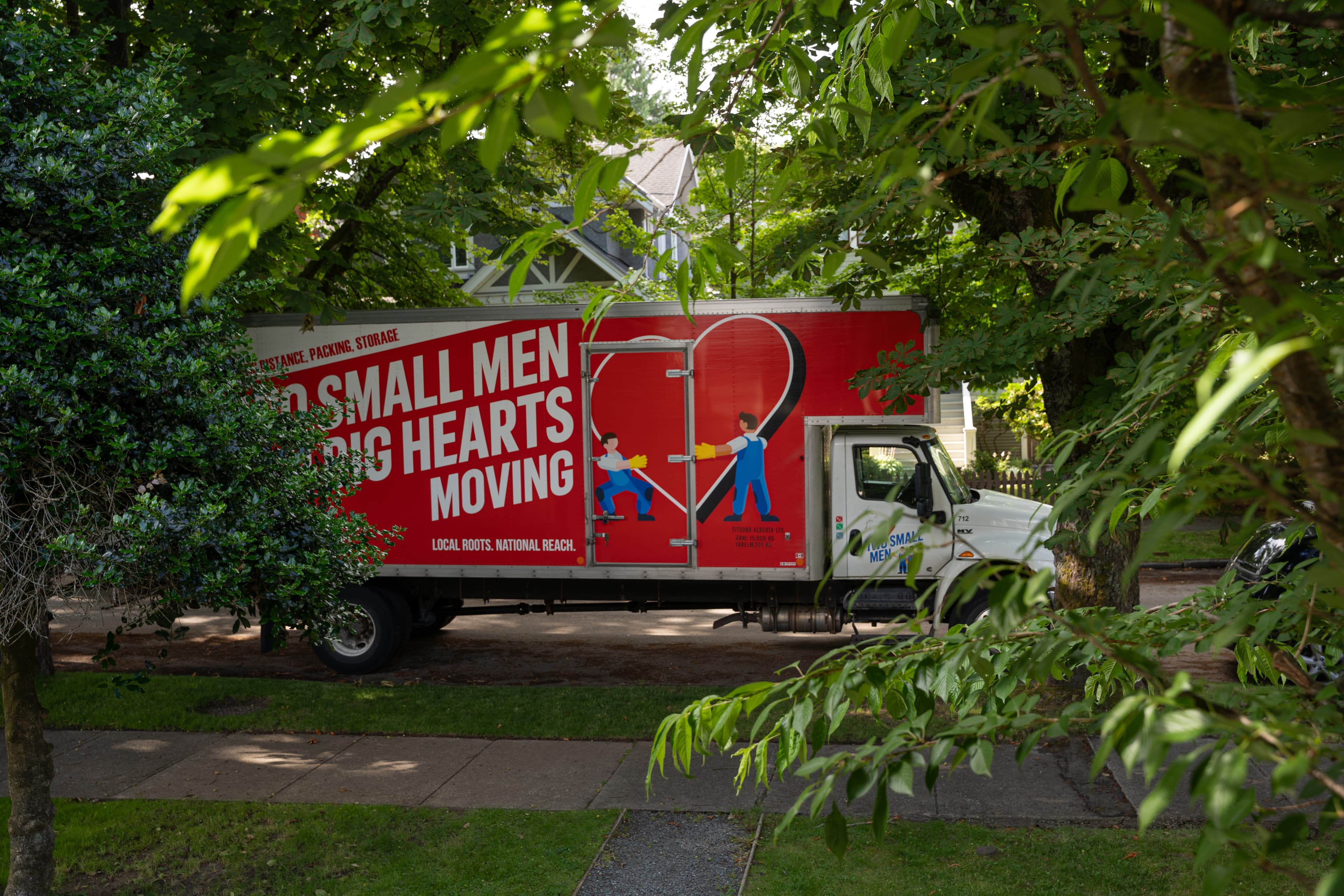 A "Two Small Men with Big Hearts Moving" truck is parked on a residential street, partially obscured by trees and greenery. The red truck features the company's logo with cartoon movers and a large heart.