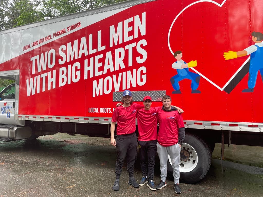 Three movers in red shirts standing in front of a Two Small Men with Big Hearts Moving truck.