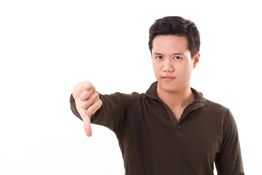 A man in a dark shirt is giving a thumbs-down gesture, with a neutral expression, standing against a plain white background.