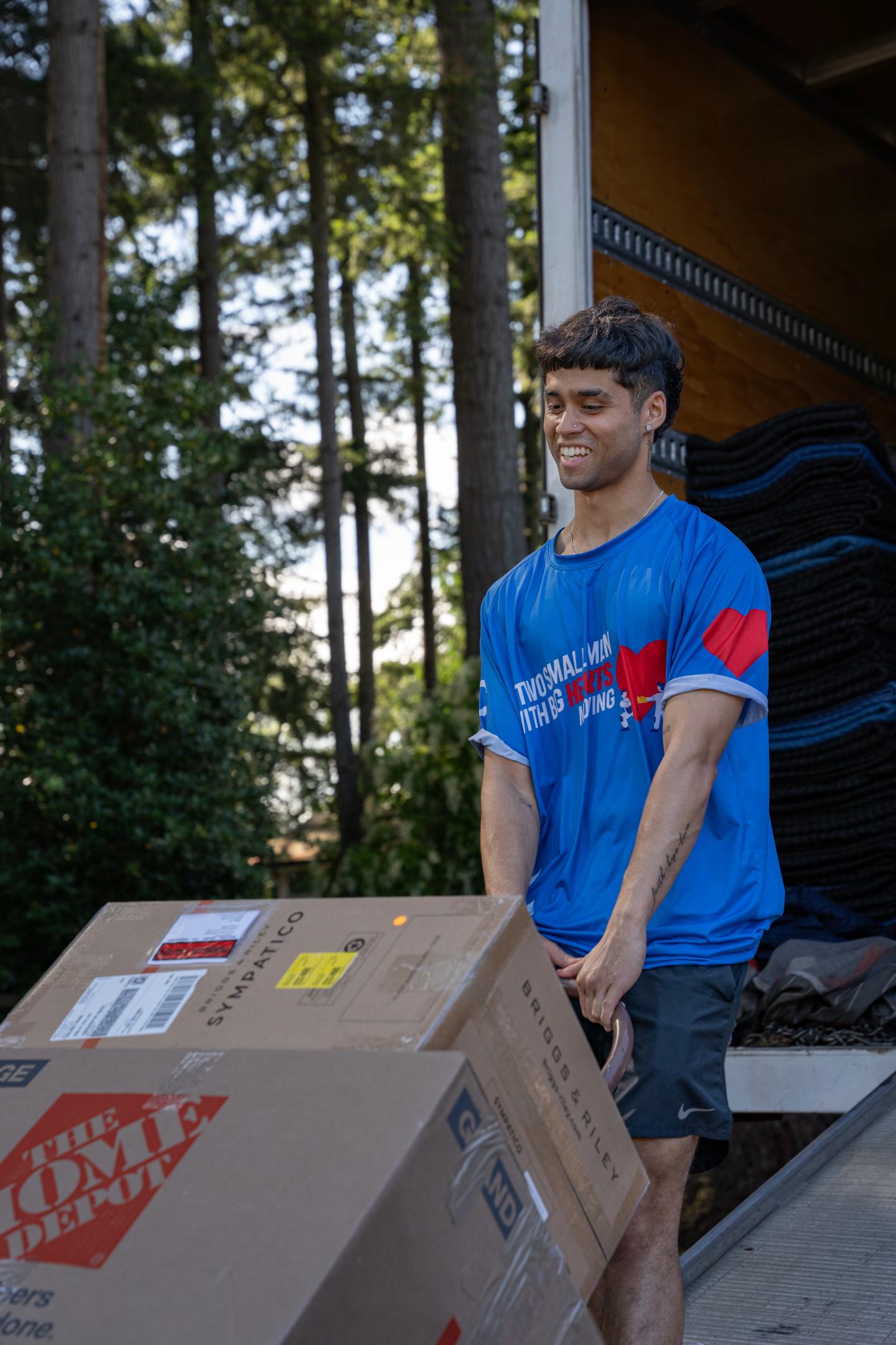 Mover in a blue "Two Small Men with Big Hearts" shirt carrying a box in front of a moving truck outdoors.