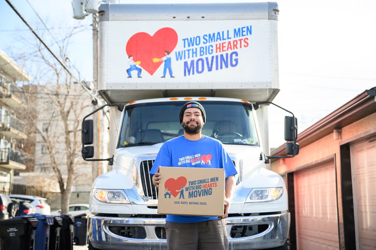 Man in a Two Small Men with Big Hearts Moving shirt holding a moving box in front of a moving truck.