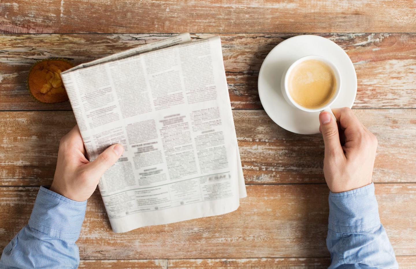 Person reading a newspaper and holding a cup of coffee at a wooden table.