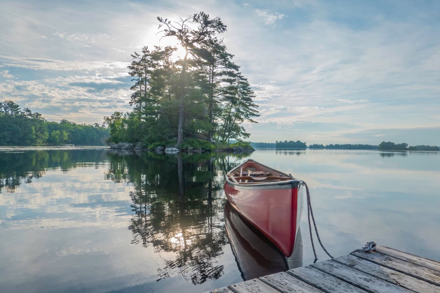 Red canoe tied to a dock on a calm lake with a small tree-covered island in Ontario.