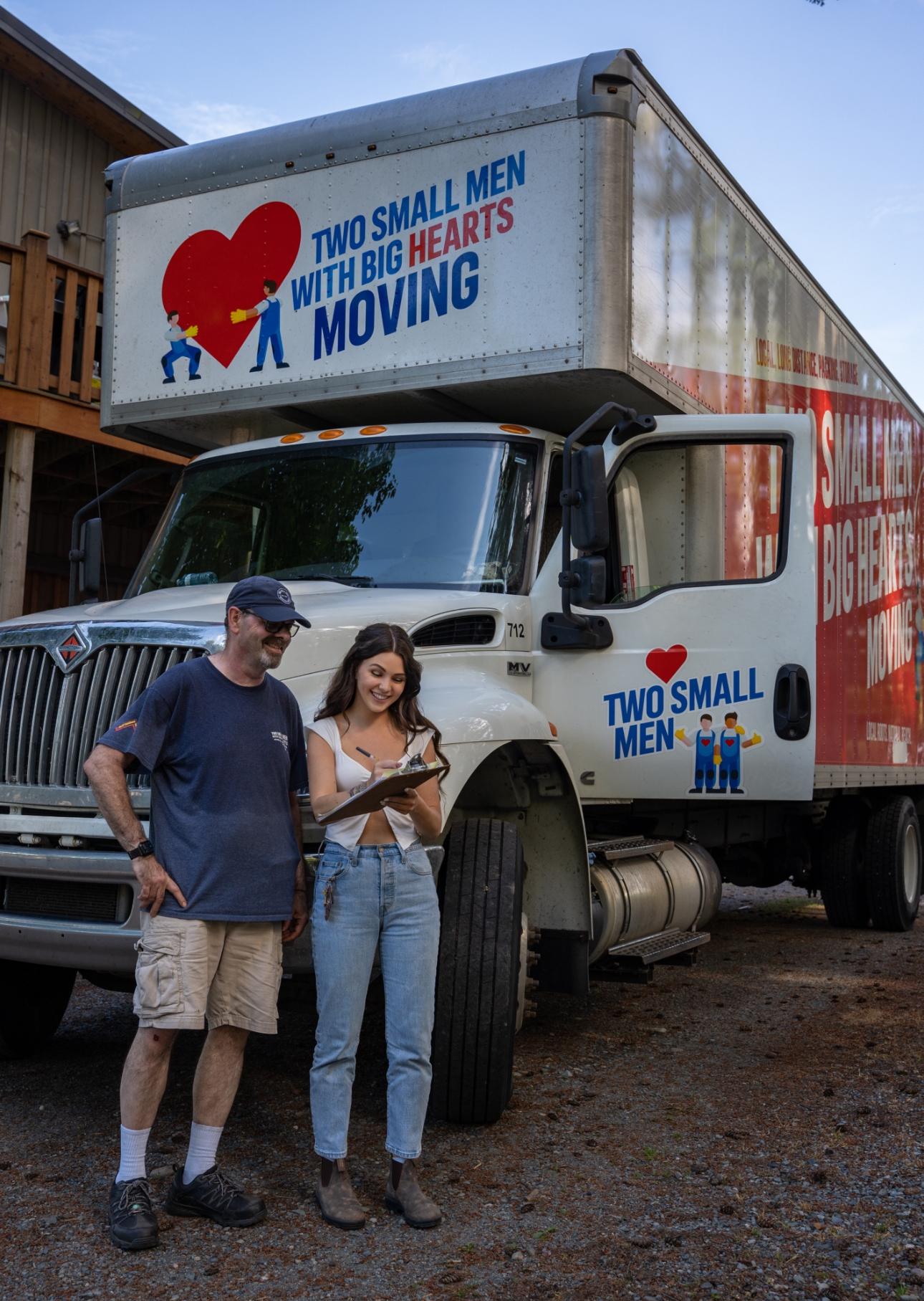 A "Two Small Men with Big Hearts Moving" truck is parked, and two people, one holding a clipboard, stand in front of it while reviewing documents. A house is partially visible in the background.