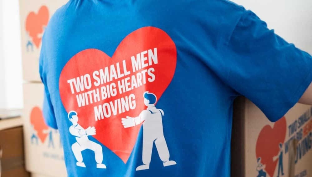 Person wearing a Two Small Men with Big Hearts Moving shirt carrying a moving box.