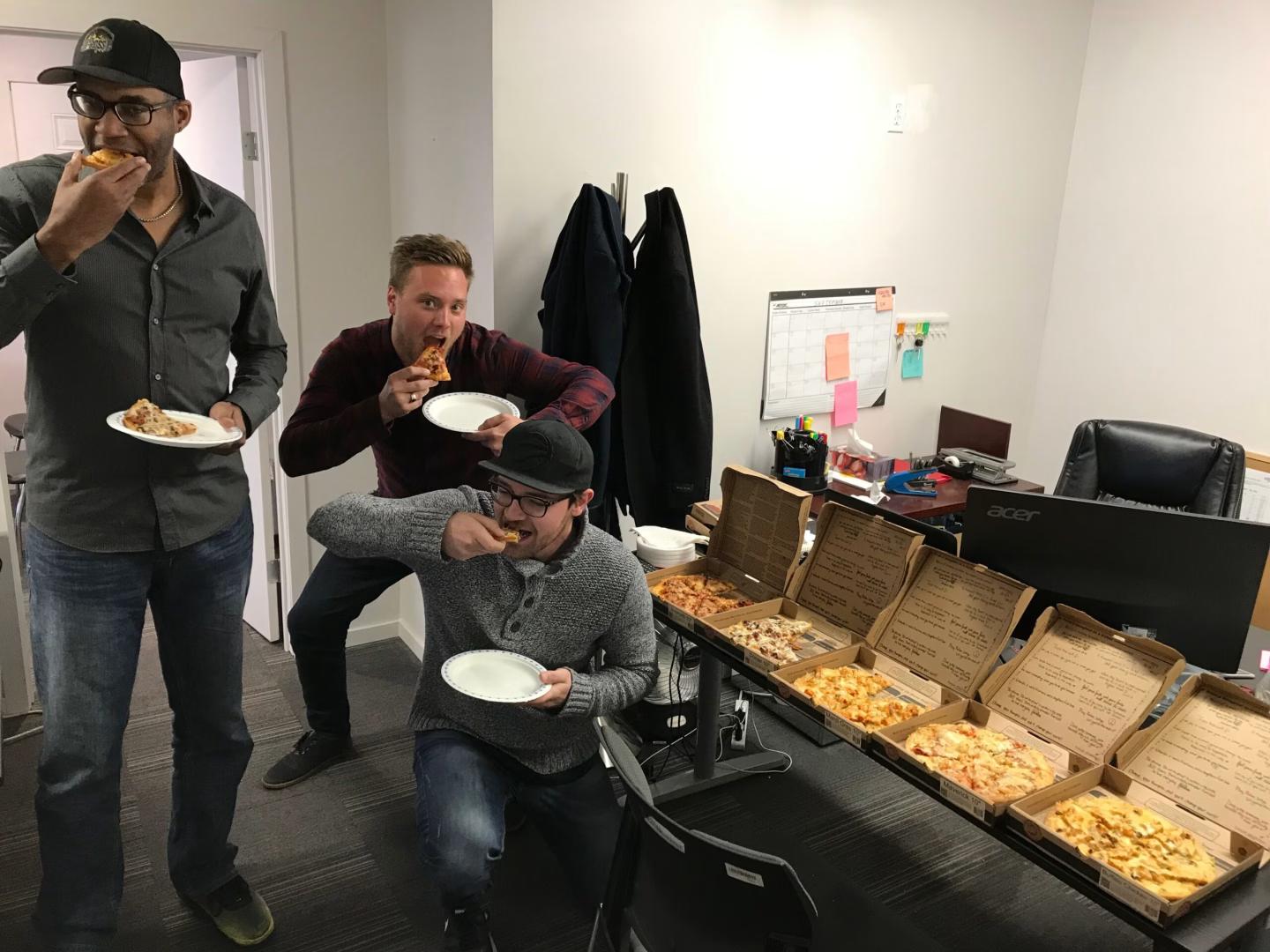 Three people are eating pizza in an office, with several open pizza boxes displayed on a table in the background. One person is standing, one is bending down, and another is kneeling while enjoying their food.