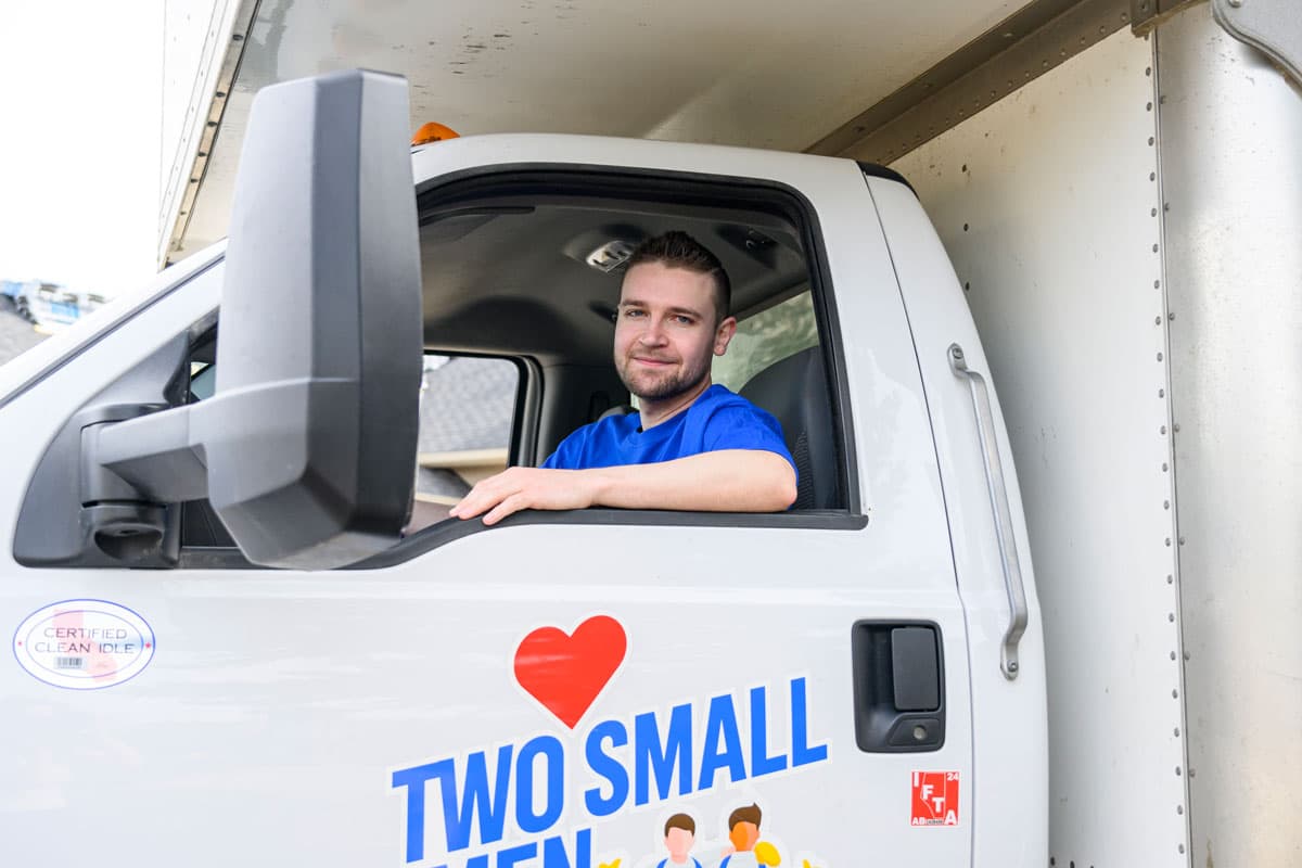 A mover sits in the driver's seat of a "Two Small Men with Big Hearts" moving truck, smiling through the open window. The company logo with a red heart is visible on the door.