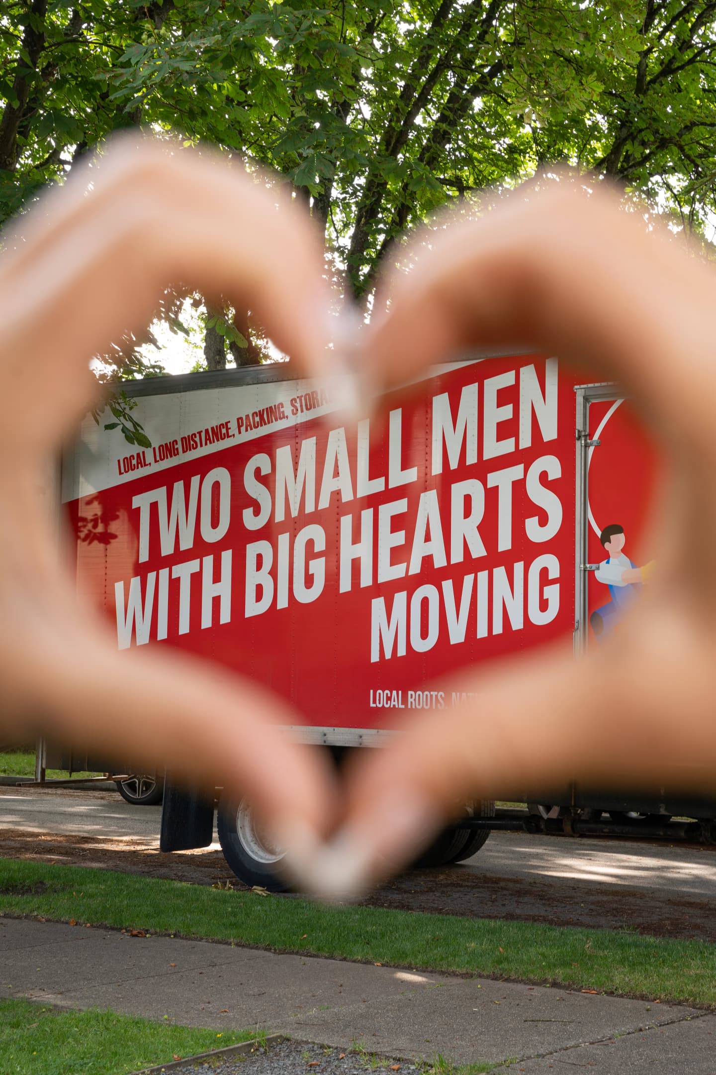 Hands forming a heart shape frame a "Two Small Men with Big Hearts Moving" truck parked outdoors with trees in the background.