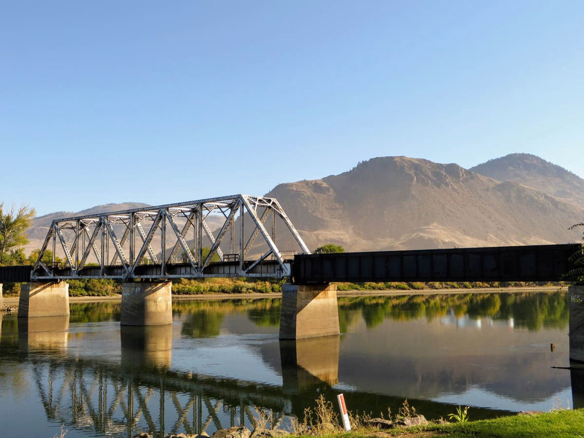 Bridge over a calm river with mountains in the background on a sunny day in Kamloops.