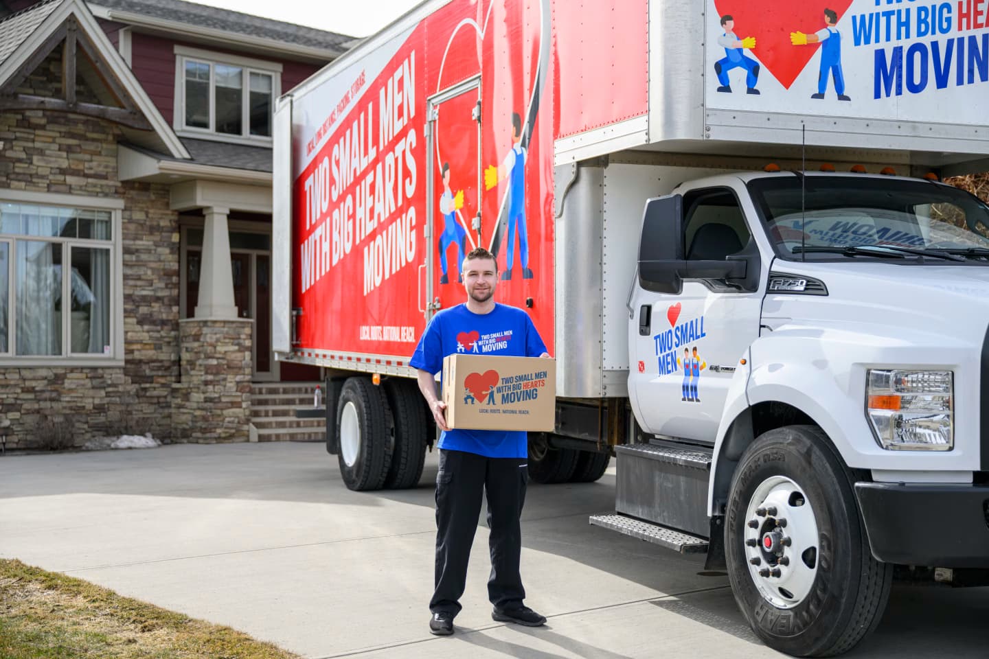 Man in a Two Small Men with Big Hearts Moving shirt holding a moving box in front of a house and moving truck.