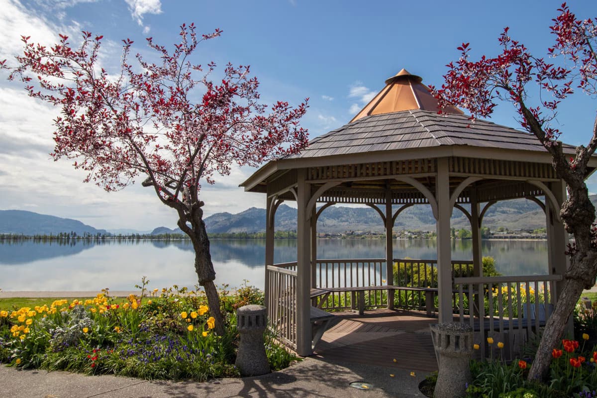 Gazebo by a lake in Osoyoos, with blooming trees and flowers, surrounded by mountains under a clear sky.