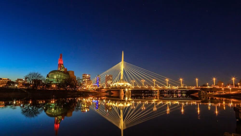 Night view of the Esplanade Riel pedestrian bridge in Winnipeg, Manitoba, illuminated with golden lights, reflecting in the river below.