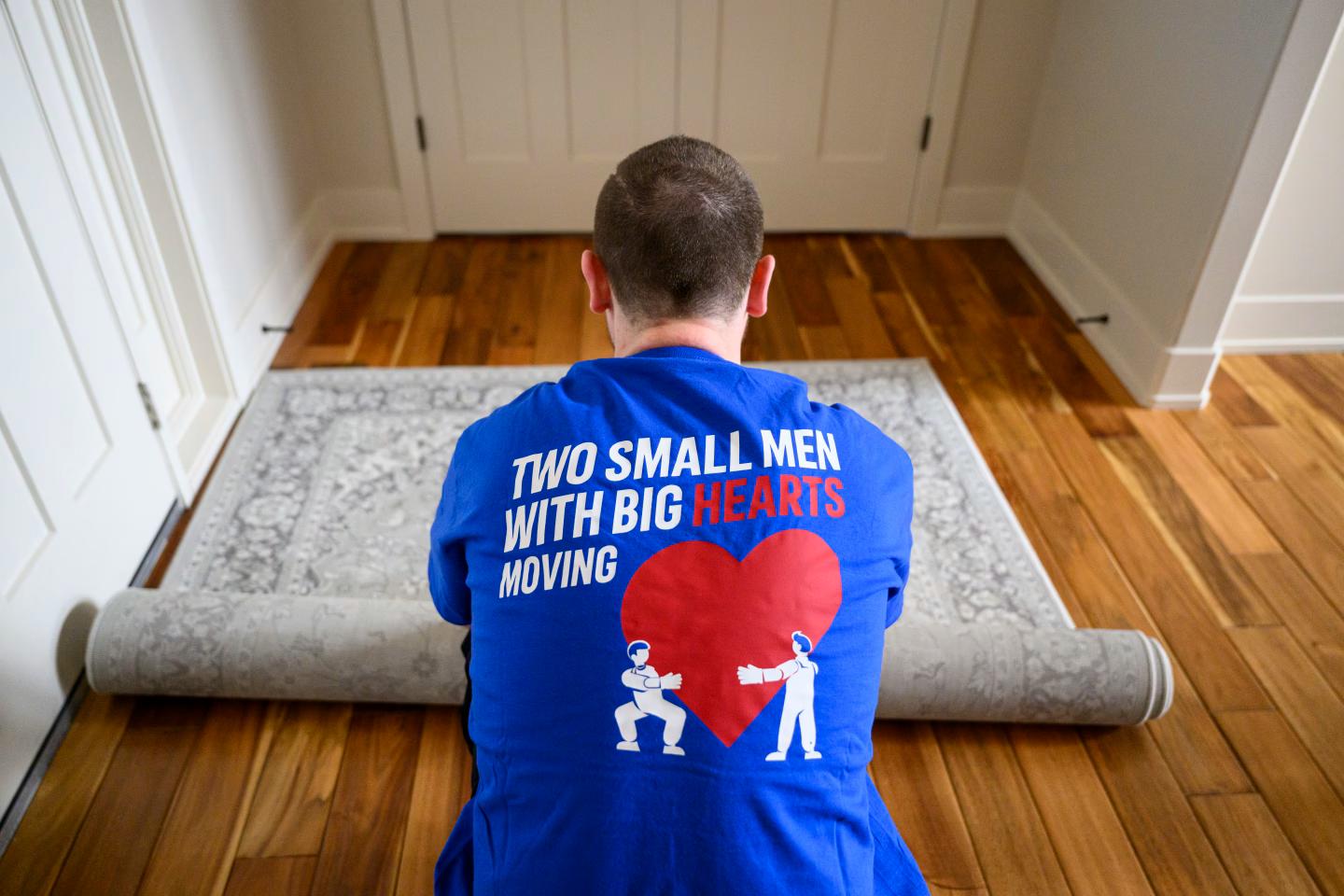 Man in a Two Small Men with Big Hearts Moving shirt rolling up a rug on a wooden floor.